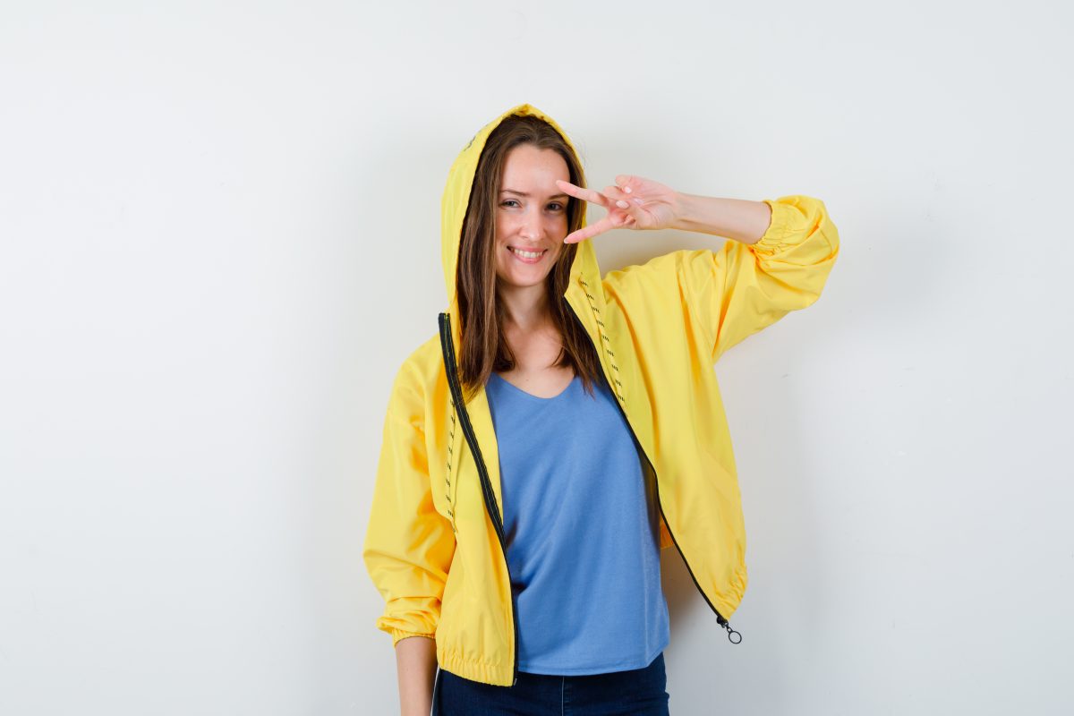 young lady showing victory gesture near eye in t shirt, jacket and looking confident , front view.